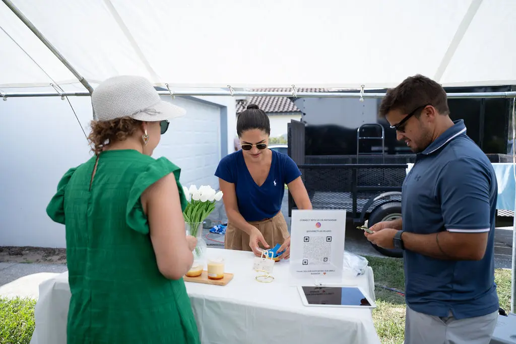A professional event vendor serving customers at an outdoor party station under a white tent, featuring a QR code sign for social media, white tulips in a vase, and small dessert cups on a table.
